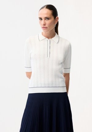 Kobieta z zaczesanymi do tyłu włosami, ubrana w biały, krótki rękaw dzianinowej koszulki polo z czarnym wykończeniem oraz granatową plisowaną spódnicę, stojąca na białym tle.