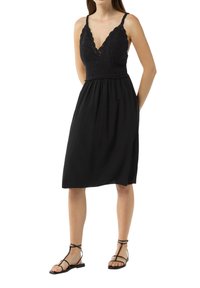 Schwarzes Kleid mit einem Häkel-Oberteil, das einen V-Ausschnitt und verstellbare Träger hat, sowie einen fließenden Rock. Getragen mit flachen schwarzen Sandalen.