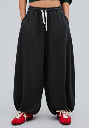 Personne portant un pantalon de survêtement noir à jambes larges avec des cordons blancs et des baskets rouges à lacets blancs, debout devant un fond uni.