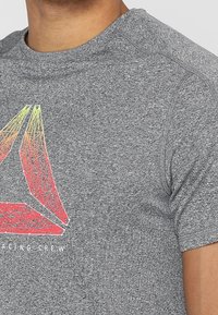 Muž ve šedém sportovním tričku s červeným a žlutým geometrickým trojúhelníkovým logem a částečným textem "RACING CREW" na hrudi.