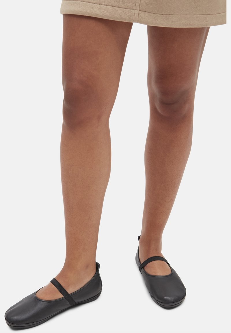 Jambes portant une jupe beige et des chaussures plates style Mary Jane noires avec des brides élastiques sur fond blanc.