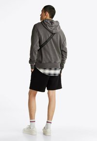 Grauer Kapuzenpullover mit strukturiertem Finish, ausgestattet mit einer Kordelzugkapuze und schwarzem Logo-Band. Kombiniert mit einem weißen Karoshirt und schwarzen Shorts.