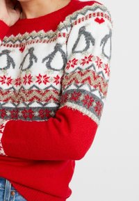 Röd stickad tröja med ett festligt mönster av grå pingviner, snöflingor och metalliska accenter. Mjuk textur med rund halsringning.