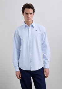 Camisa azul claro de botones con mangas largas, que presenta un sutil patrón de rayas y un pequeño logo bordado en el pecho.