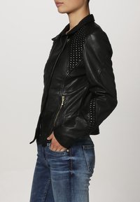 Giacca di pelle nera con dettagli in borchie, chiusura zip asimmetrica e design aderente. Abbinata a jeans blu, mette in risalto una silhouette elegante.