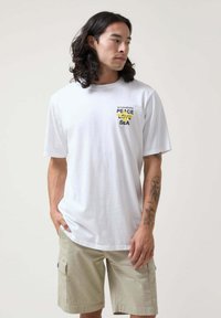 Camiseta blanca de algodón con mangas cortas, que presenta un estampado gráfico que incluye "PAZ" y "MAR" en amarillo y negro. Combinada con pantalones cortos color caqui claro.