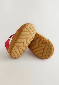 Zapatos infantiles rojos con un logo blanco, fabricados en un tejido suave y con suelas de goma que presentan un patrón marrón texturizado y bordes redondeados.