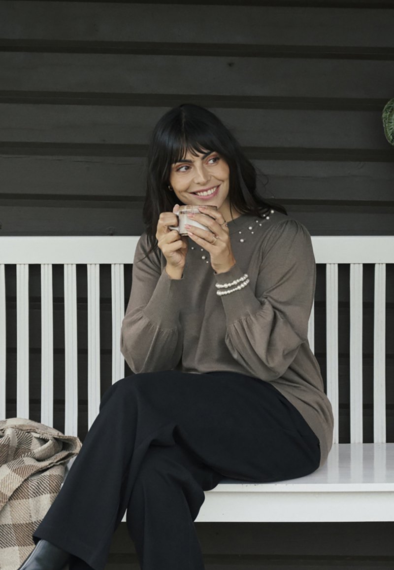 Hnedý sveter s perlovými detailmi na výstrihu, dlhé rukávy so zbieranými manžetami, v kombinácii s čiernymi nohavicami, drží pohár.