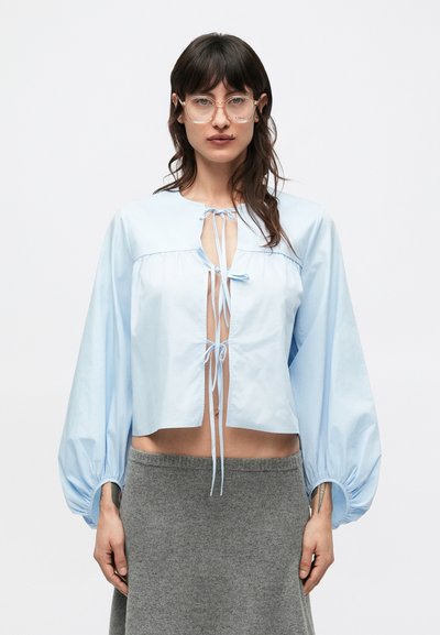 Light blue cropped shirt in lightweight fabric, featuring puff sleeves, tie closures at the front, and a relaxed fit. Paired with a gray skirt.