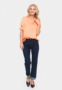 Satinaste oranžno bluza s napihnjenimi rokavi in ovratnikom, v kombinaciji s temno modrimi kavbojkami, zavihanimi na manšetah, in rjavimi špicastimi petami.