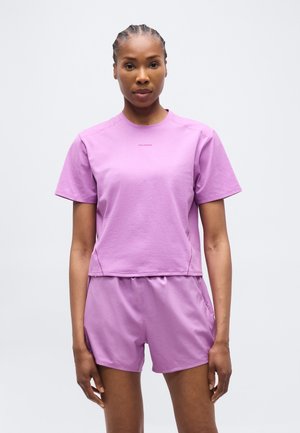 Γυναίκα με πλεξιές στα μαλλιά, φορώντας ένα ολόιδιο σετ από λιλά κοντομάνικη μπλούζα και σορτς, που στέκεται μπροστά σε ένα απλό φωτεινό φόντο.