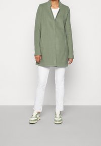 Persona che indossa un lungo cappotto verde chiaro, pantaloni bianchi e sneakers bianche e verde chiaro, in piedi su un pavimento grigio chiaro contro un muro bianco.