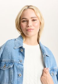 Jasnoniebieska kurtka dżinsowa z guzikami, połączona z białym swetrem w warkocze. Modelka ma proste blond włosy i trzyma kurtkę.