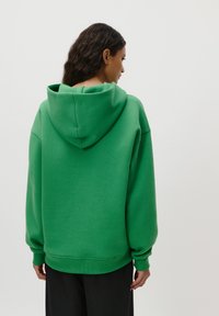 Kvinne med langt mørkt krøllet hår iført en oversized grønn hettegenser og svarte bukser, vendt bort fra en ensfarget hvit bakgrunn.