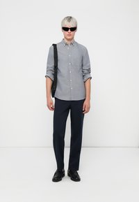 Grijze button-up overhemd met opgerolde mouwen, marineblauwe op maat gemaakte broek, zwarte zonnebril en zwarte schoenen, staande tegen een witte achtergrond.