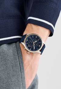 Navy watch with a gold stainless steel case, textured navy strap, and dark blue dial featuring silver accents and three subdials.