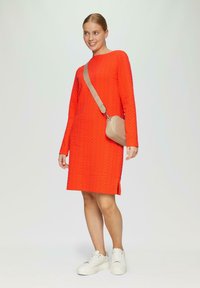 Rotes strukturiertes Strickkleid mit langen Ärmeln, seitlichem Schlitz und einem lässigen Schnitt. Kombiniert mit einer beige Umhängetasche und weißen Plateauschuhen.