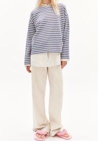 Niebiesko-beżowa pasiasta bluzka z długim rękawem, nałożona na białą teksturowaną koszulę, zestawiona z jasnobeżowymi spodniami o szerokich nogawkach i różowymi sandałami.