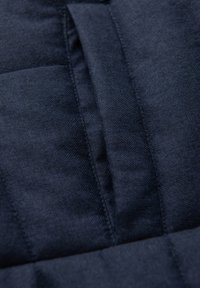 Detailní záběr tmavě modré látky se vzorem rybí kosti a středovým švem s detaily složeného šití.