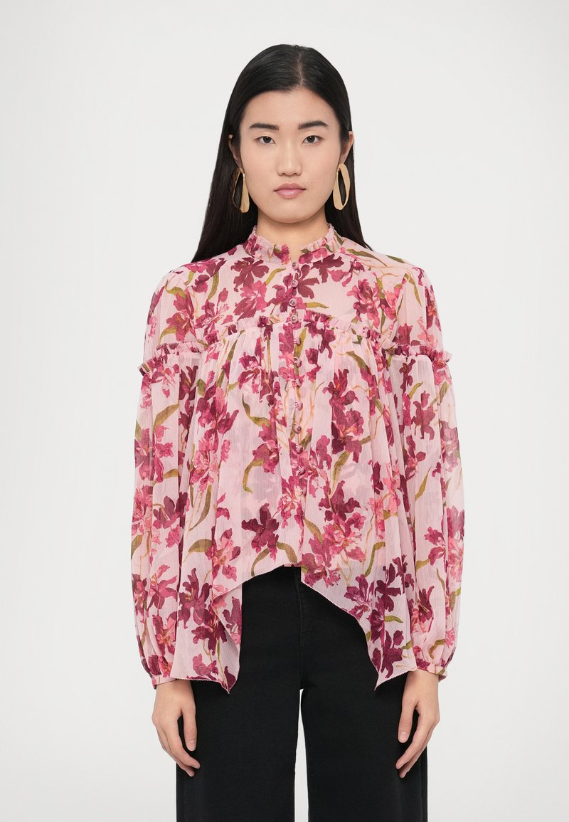 Blouse fleurie en tissu léger et transparent, ornée de fleurs roses, rouges et vertes. Col montant, boutonnage sur le devant, et design asymétrique de l'ourlet.