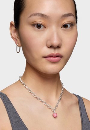 Primer plano de una persona que lleva un collar de cadena plateada con un colgante de corazón rosa y un pendiente de aro plateado, sobre un fondo liso.