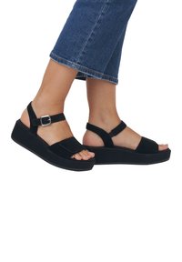 Černé semišové platformové sandály se širokým prstovým páskem a nastavovacími popruhy kolem kotníku. Mají pevnou černou podrážku a minimalistický design.