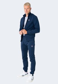 Chaqueta de cremallera azul marino con cuello alto, combinada con pantalones jogger a juego. Ambos cuentan con un logo en el pecho y la cadera. Zapatillas blancas completan el conjunto.