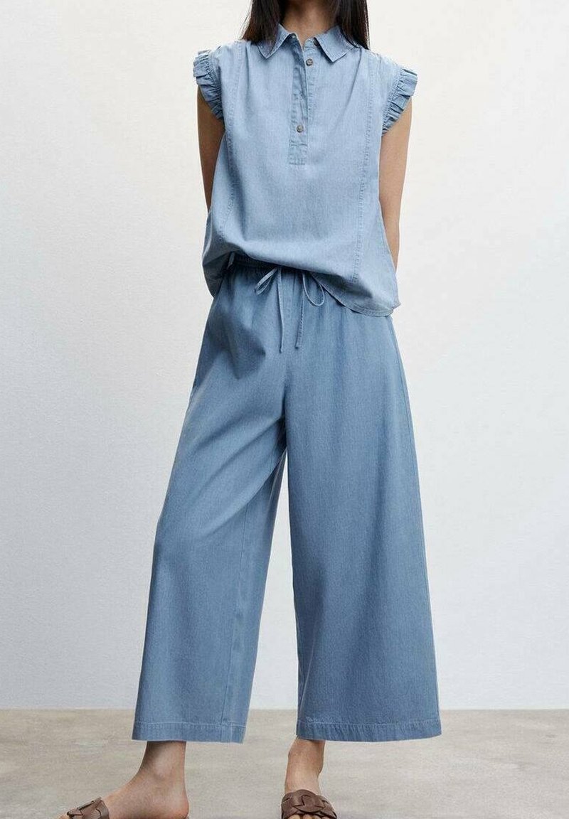 Femme portant un haut en denim léger bleu clair sans manches avec des épaules froncées et un pantalon large assorti avec taille à cordon et des sandales marron.