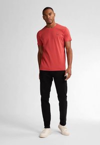 Camiseta de algodón color carmesí con cuello redondo, mangas cortas y un pequeño logo en el pecho izquierdo, combinada con pantalones negros y zapatillas de color claro.