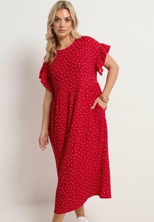 Kvinde iført en rød midikjole med hvide polkaprikker, flæser på korte ærmer og sidelommer, stående foran en ensfarvet baggrund.