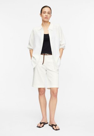 Kobieta stoi z rękami w kieszeniach, ubrana w białą koszulę na guziki, czarny top, białe krótkie spodenki do kolan, pleciony pasek i czarne sandały.