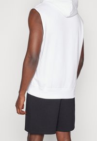 Persona che indossa un hoodie bianco senza maniche e pantaloni corti neri, mostrata di spalle, in piedi su uno sfondo chiaro e semplice.