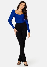 Top blu lungo a maniche arricciate con scollo quadrato, abbinato a pantaloni neri a vita alta a campana e sandali neri platform. Sfondo bianco.