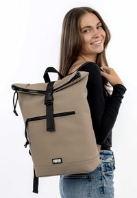 Zaino roll-top impermeabile beige con dettagli neri della cerniera, spallacci regolabili e tasca frontale. Tessuto liscio con un design minimalista.