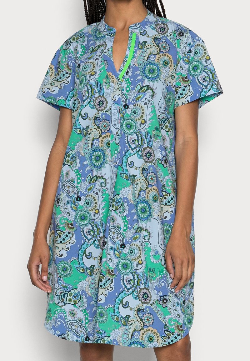 Femme portant une robe courte à manches courtes, longueur genou, avec un motif floral paisley bleu, vert et blanc et un col en V avec bordure vert néon.