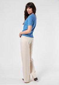 Vaaleansininen lyhythihainen neulepaita, jossa on teksturoitu timanttikuosi, yhdistettynä beigeihin leveälahkeisiin housuihin ja ruskeisiin korkokenkiin.