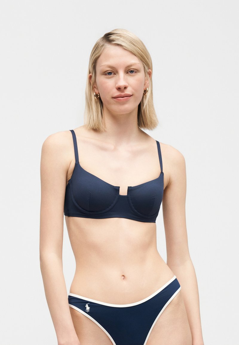 Γυναίκα με ίσια ξανθά μαλλιά, ντυμένη με μπλε σουτιέν και μαγιό μπικίνι με άσπρη μπορντούρα, σε απλό φόντο.