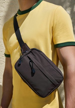 Persoană care poartă un tricou galben cu tiv verde și o geantă neagră purtată în diagonală, cu două compartimente cu fermoar, în lumină exterioară.