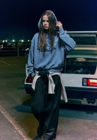 Világoskék kapucnis pulóver "Stranger Things" felirattal, laza fekete nadrággal párosítva. Fehér pulóver a derekán megkötve, hosszú barna haj. Éjjeli környezet.