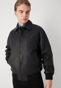 Melnas ādas bumbieriņa jaka ar pogu aizdari, ribotām piedurknēm un saliektu apkakli. Uz krūtīm ir izspiests "HUGO" logo.