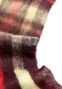 Wełniany szalik w odcieniach czerwieni i żółci, o miękkiej, puszystej strukturze i kratkowanym wzorze. Czarna metka z logo znajduje się blisko krawędzi.