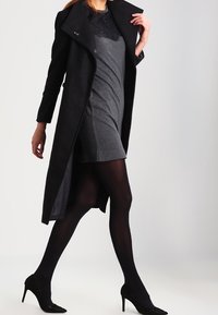 Lungo cappotto nero con colletto strutturato, indossato sopra un vestito corto grigio. Collant neri e scarpe con tacco alto completano l'outfit. Tessuto liscio.