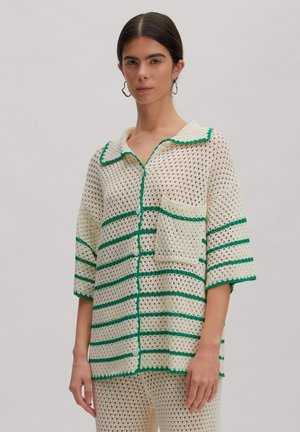 Vrouw draagt een crème gehaakt overhemd en broek met groene strepen, korte mouwen, kraag, knopen en voorzak, staand tegen een effen achtergrond.