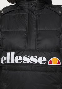 Chaqueta de plumas negra con un alto cuello, que cuenta con un gran logo blanco y un acento en amarillo y rojo en el detalle del logo en la parte inferior frontal.