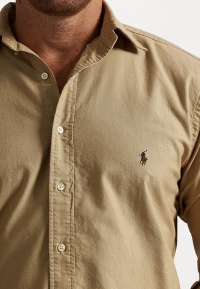 Barna gombos ing pamutból, klasszikus gallérral, elülső gombolással és egy kis hímzett logóval a bal mellrészen.