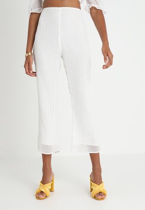 Pantalons crops plissés blancs avec un léger éclat, présentant une coupe droite et une longueur cheville. Associés à des sandales à talons carrés jaunes.