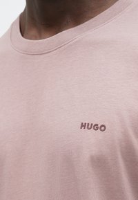 Tricou roz din bumbac, cu un guler rotund clasic și un mic logo „HUGO” în relief, într-o nuanță mai închisă, pe partea stângă a pieptului.