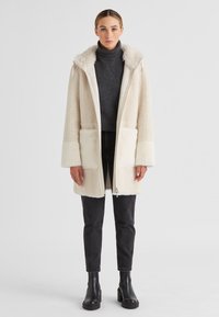 Frau in einem cremefarbenen strukturierten Kapuzenmantel über einem dunkelgrauen Rollkragenpullover, schwarzen Jeans und schwarzen Stiefeletten, steht vor einem weißen Hintergrund.