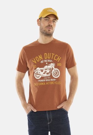 Homme portant un t-shirt orange Von Dutch avec un graphique de moto vintage et une casquette jaune, se tenant avec les mains dans les poches.