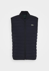 Tmavě modrá vesta bez rukávů s prošívaným vzorem, předním zipem, vysokým límcem a malým zeleným logem Lacoste na hrudi.